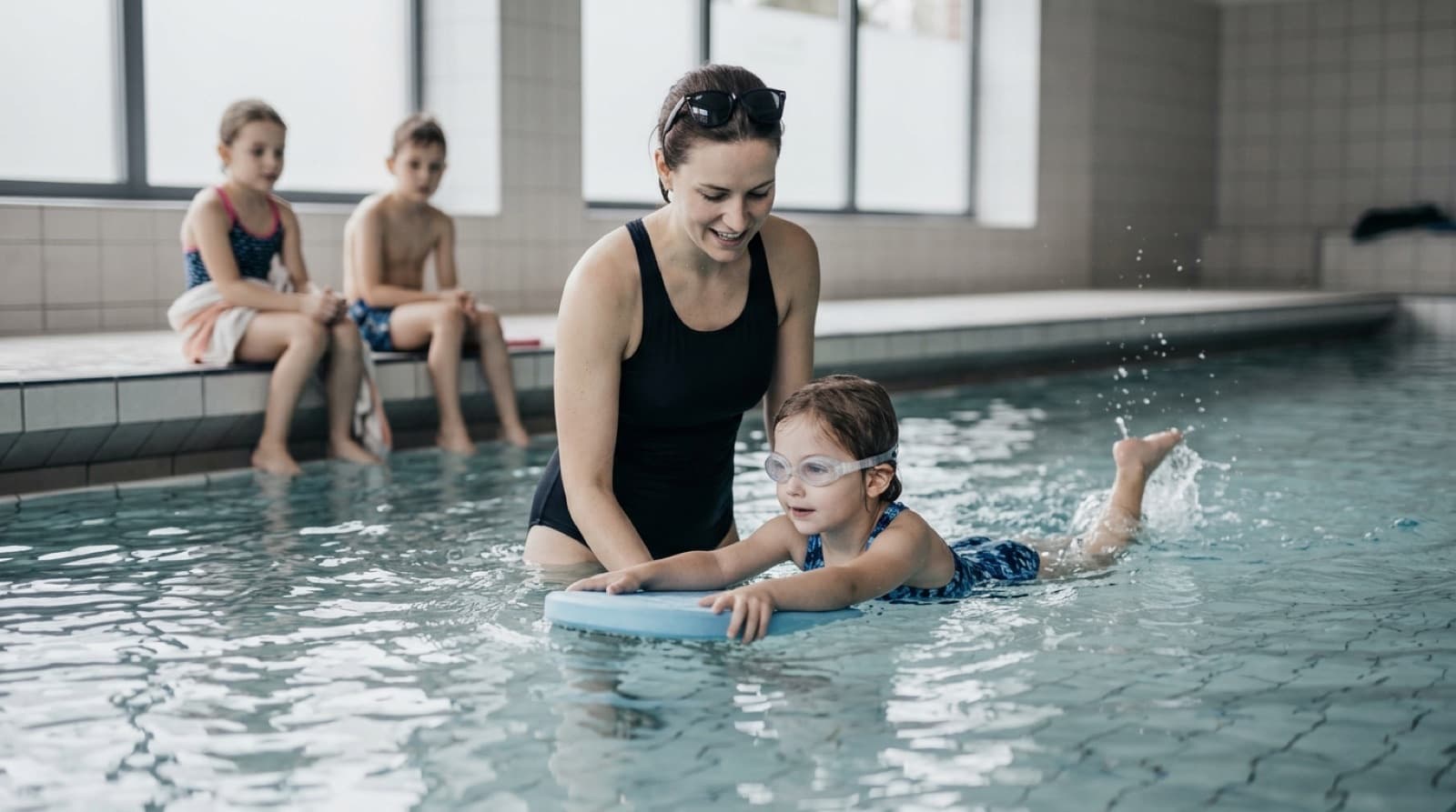 Eine Schwimmlehrerin steht im Lehrschwimmbecken und begleitet ein Mädchen mit Schwimmbrille, das sich mit einem Schwimmbrett nach vorn gleiten lässt, zwei Kinder warten am Beckenrand